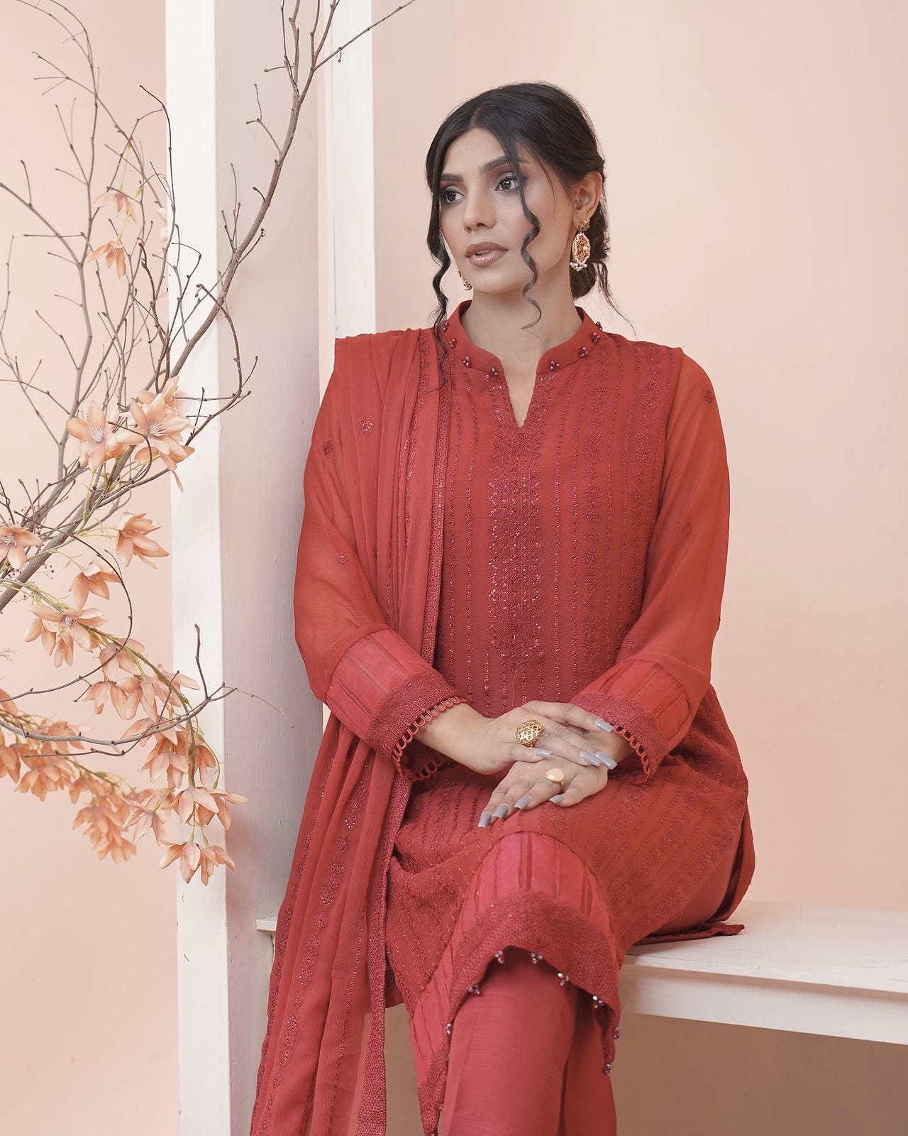 A woman sits beside a vase of dried branches in a peach-lit room, wearing a coral-red kurta set.
