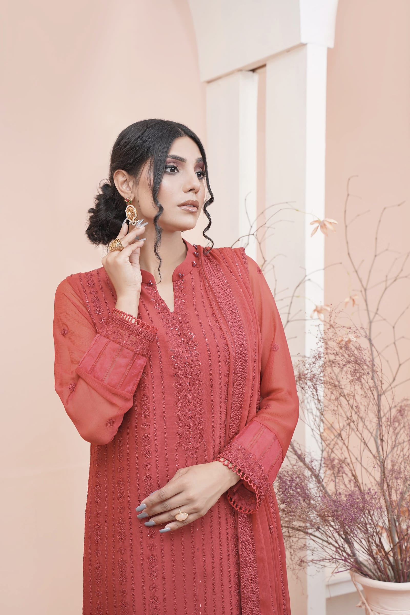 A woman is close in a peach-lit room, wearing a coral-red kurta set.