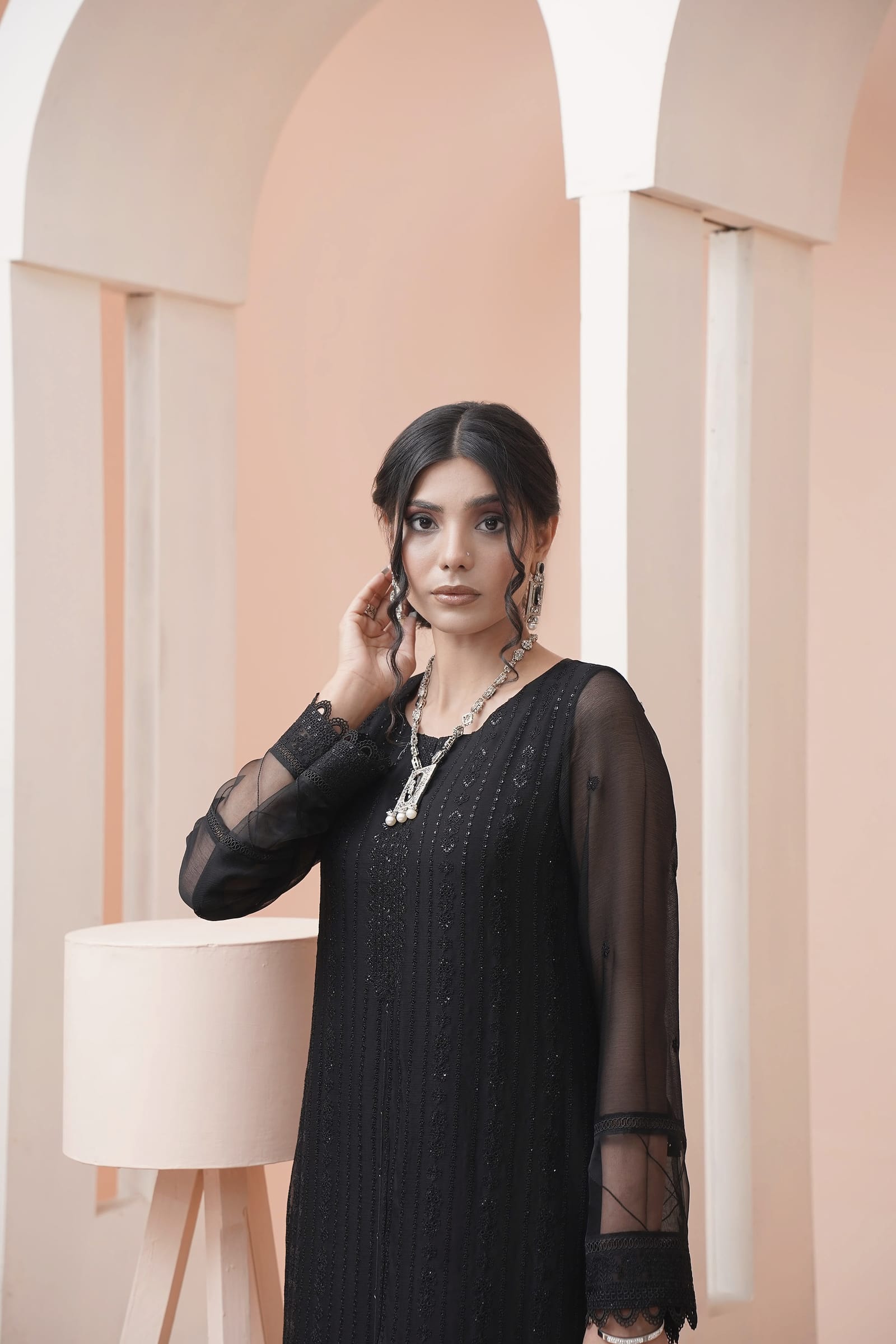 A woman is close in a peach-lit room, wearing a black sheer kurta set with an embroidered placket and a black dupatta.