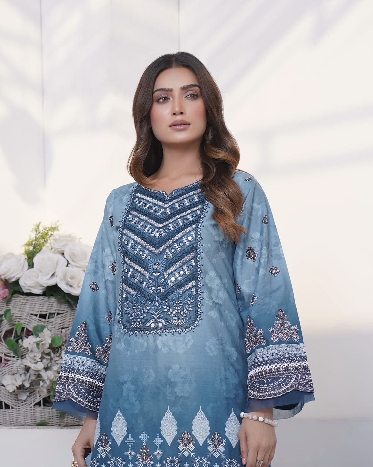 A woman in a sky-blue ombre kurta set with a navy chevron yoke, shown to the hips, with white roses and a framed portrait behind her.