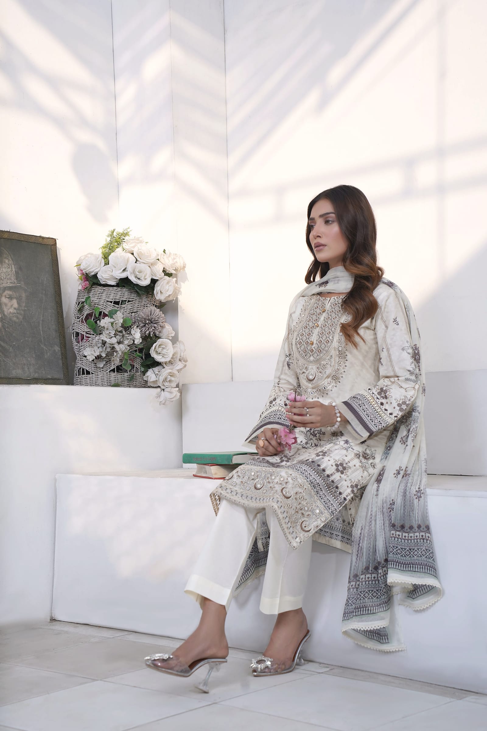 A woman sits on a white step in a white room, wearing an ivory kurta set with a grey-black print and a pale ombre dupatta.