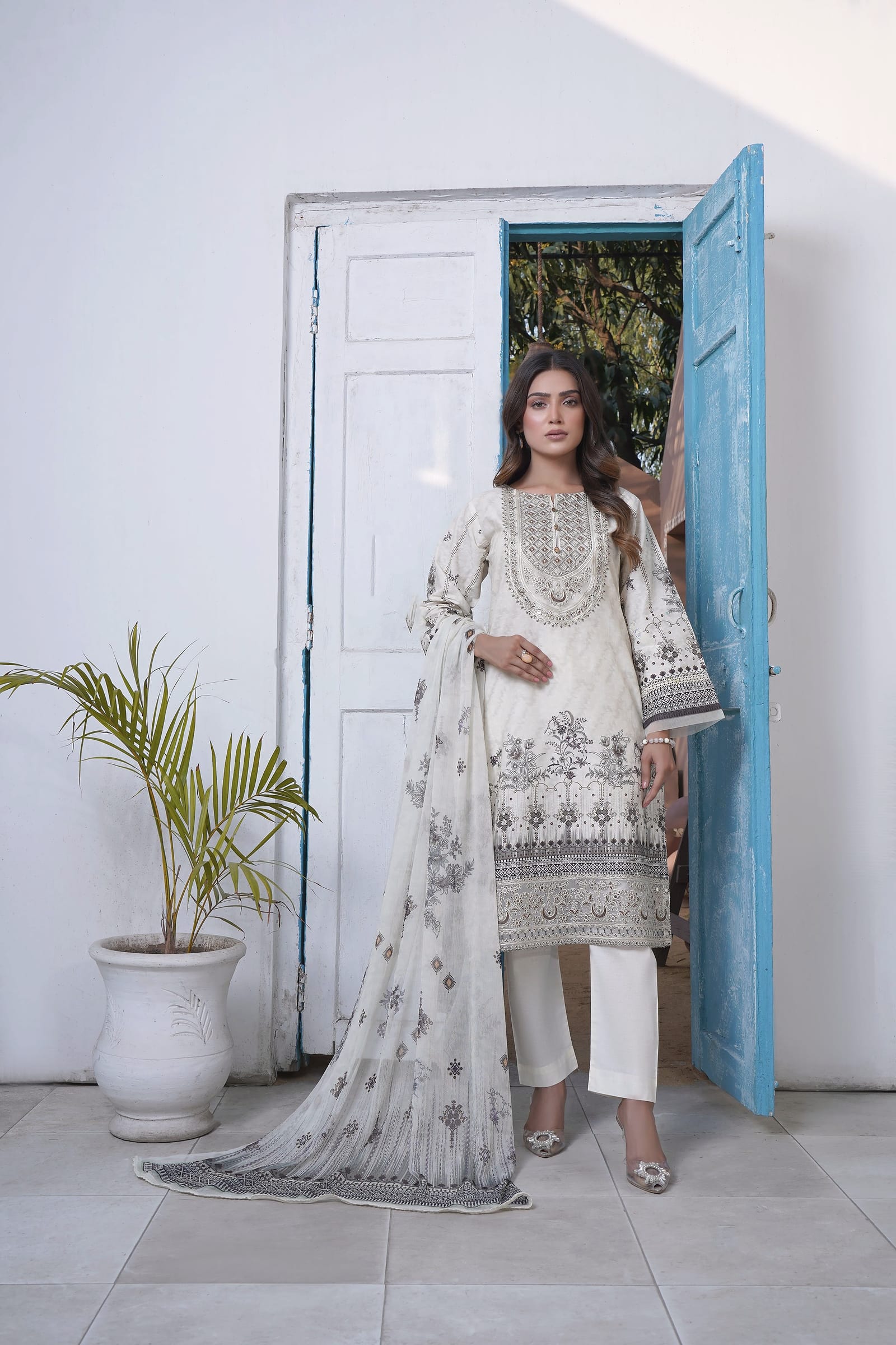 A woman stands full length in front of an open blue door, wearing an ivory kurta set with a grey-black print and a pale ombre dupatta.