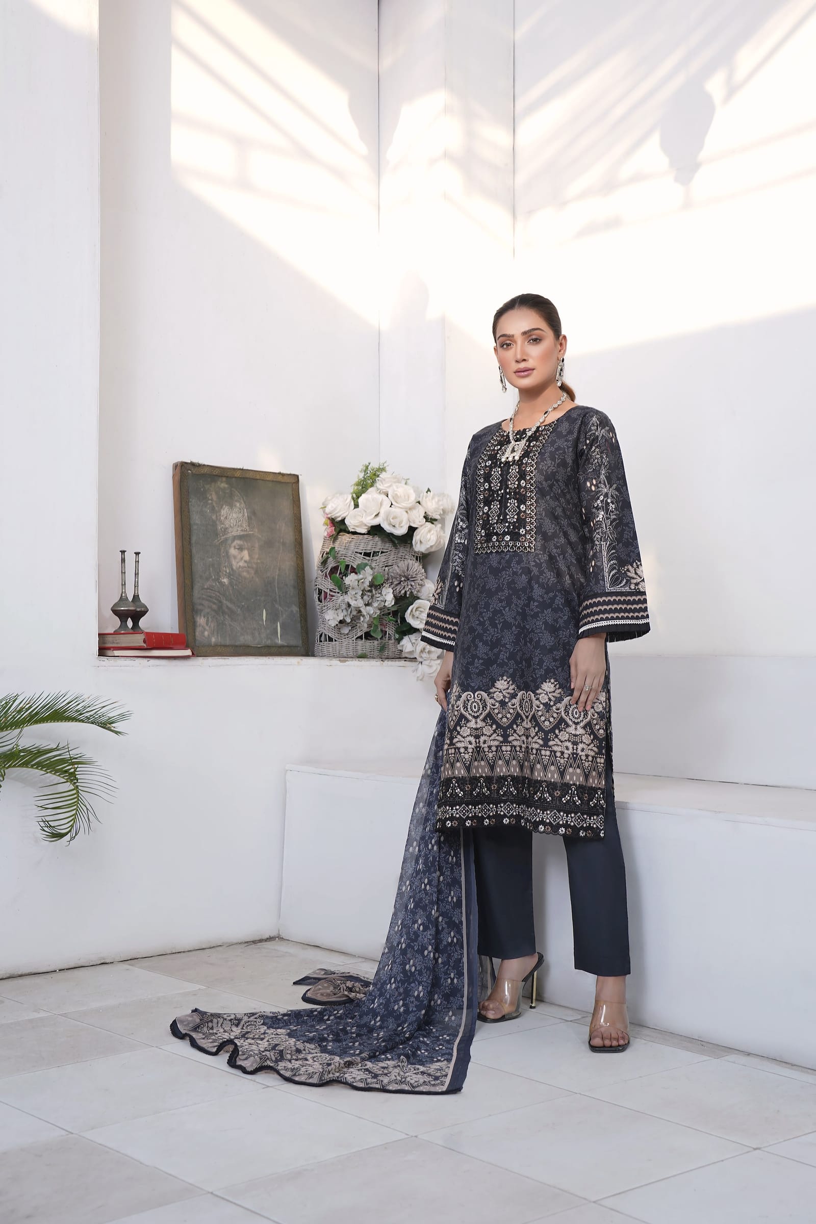 A woman stands full length in a white room, wearing a slate-navy kurta set with a black embroidered yoke.