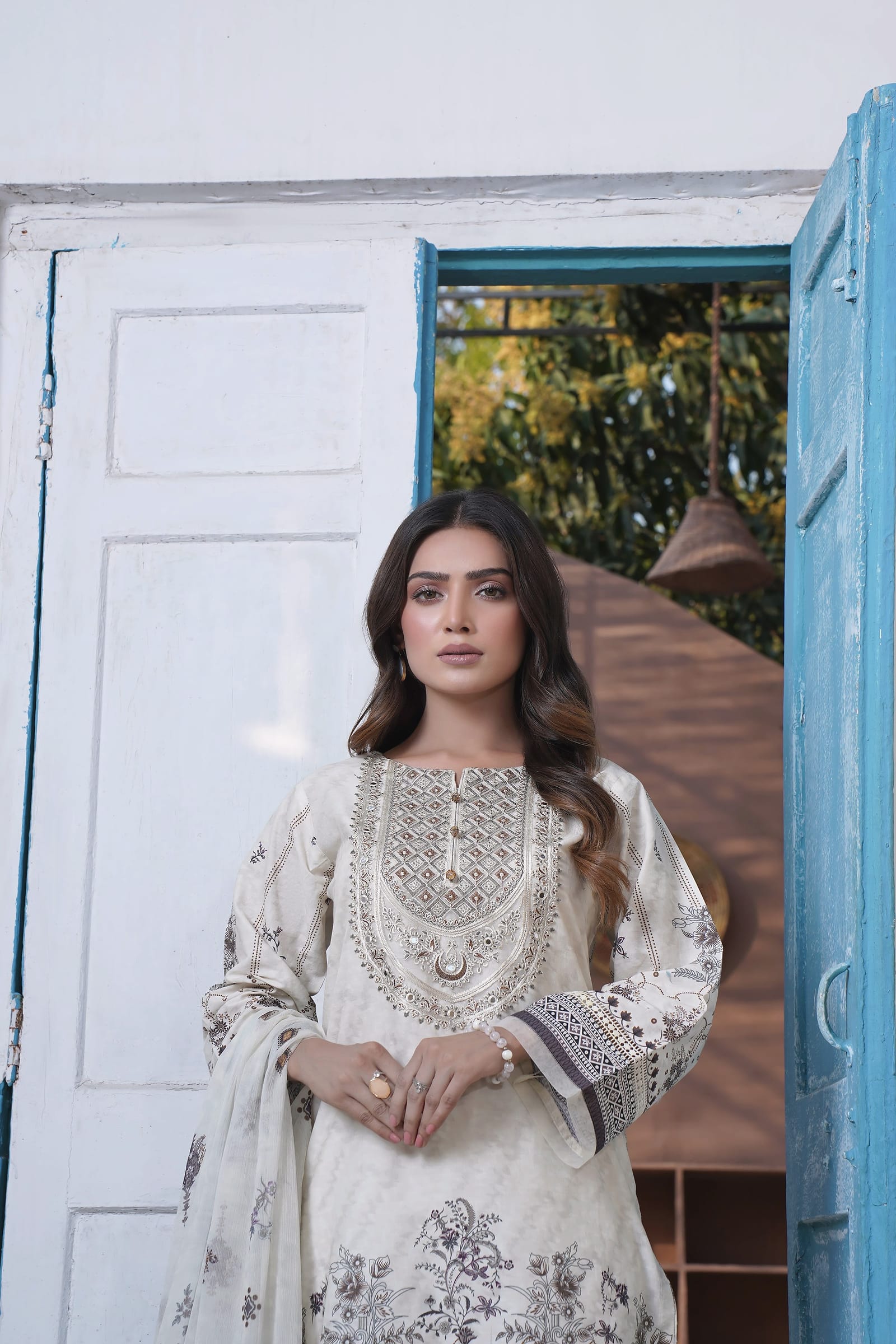 A woman stands three-quarter length in front of an open blue door, wearing an ivory kurta set with a grey-black print and a pale ombre dupatta.