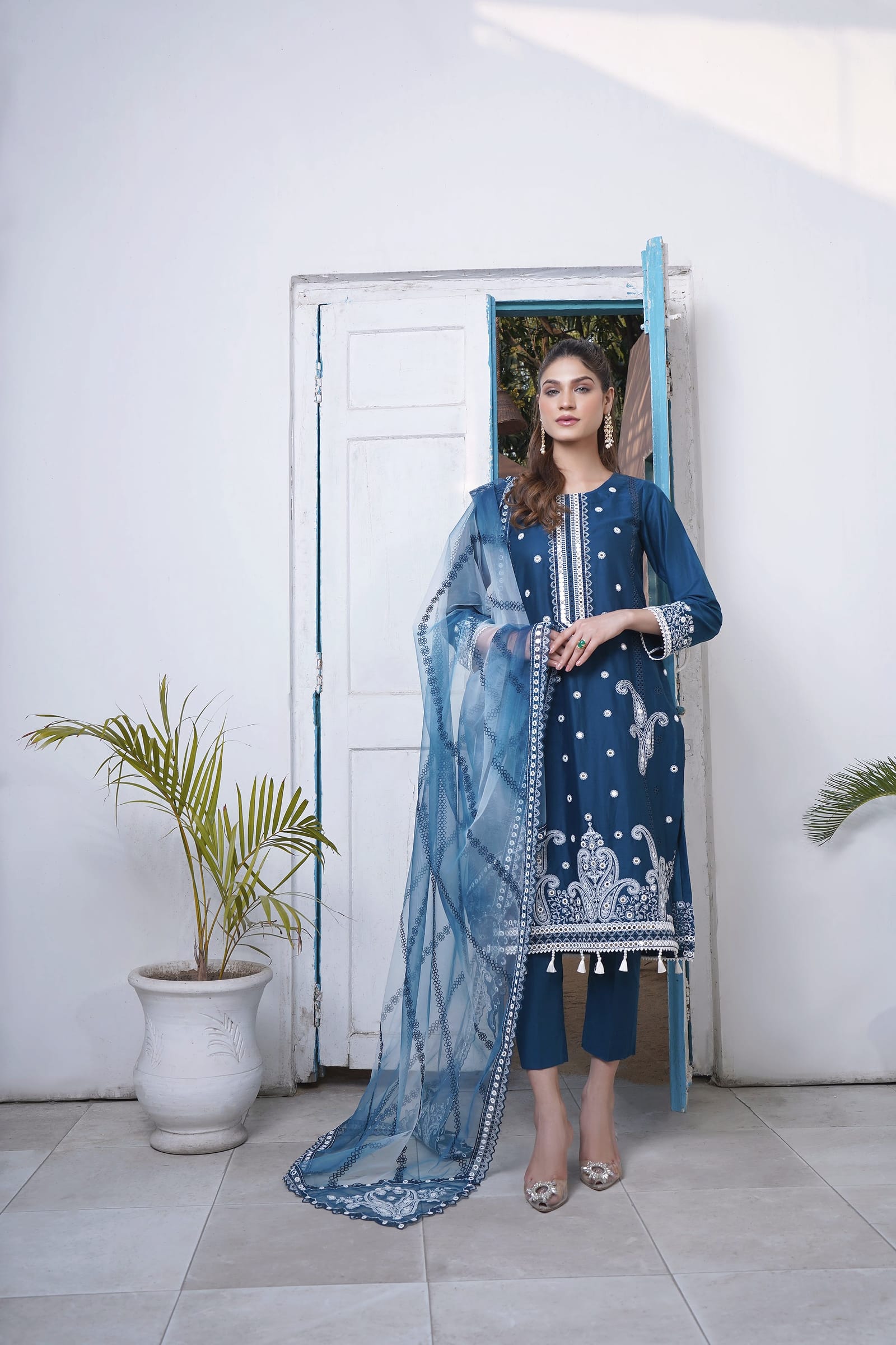A woman stands full length in front of an open blue door, wearing a peacock teal-blue kurta set with white paisley embroidery and a pale blue net dupatta.