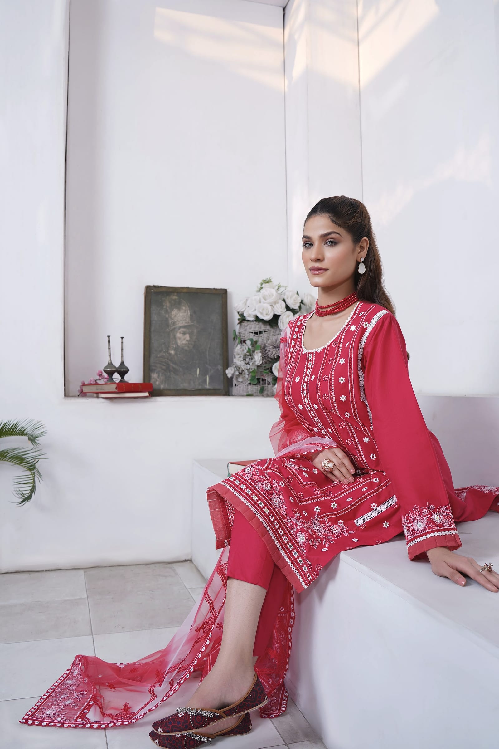 A woman sits on a white step, wearing a raspberry-red kurta set with white embroidery and a red net dupatta.