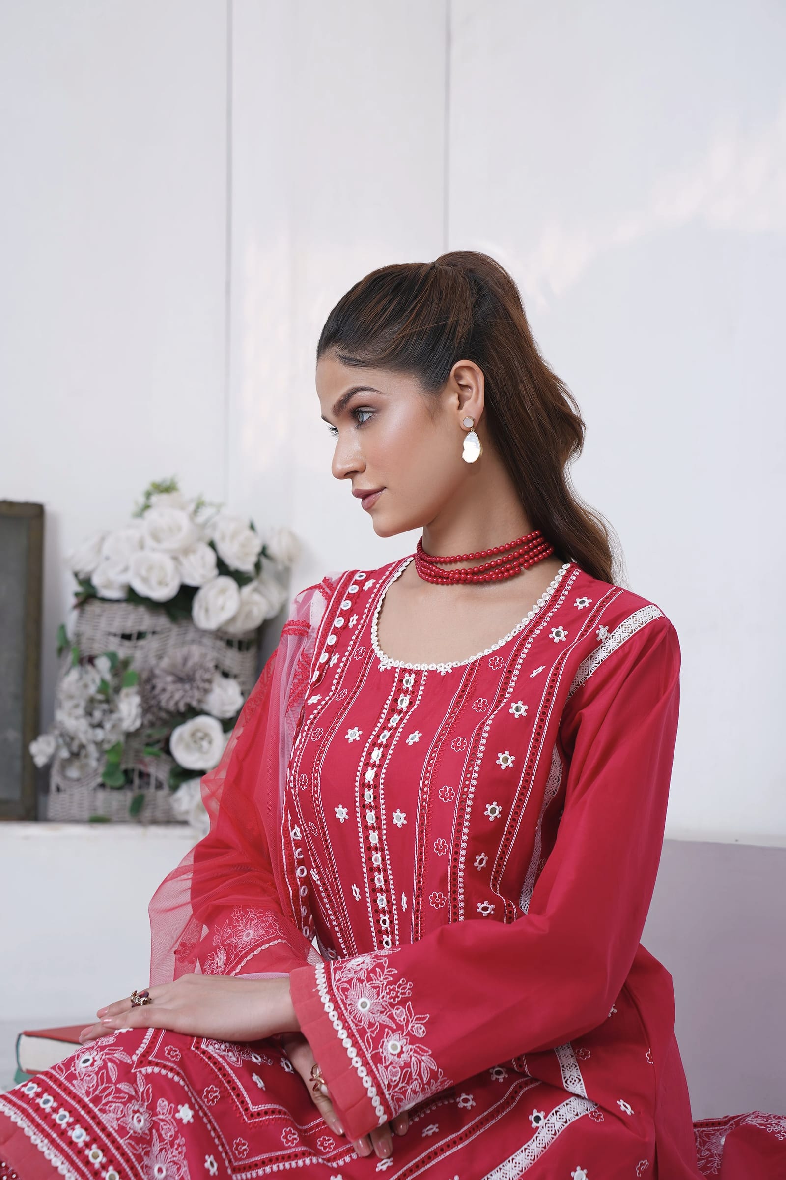 A woman is close in front of white roses, wearing a raspberry-red kurta set with white embroidery and a red net dupatta.
