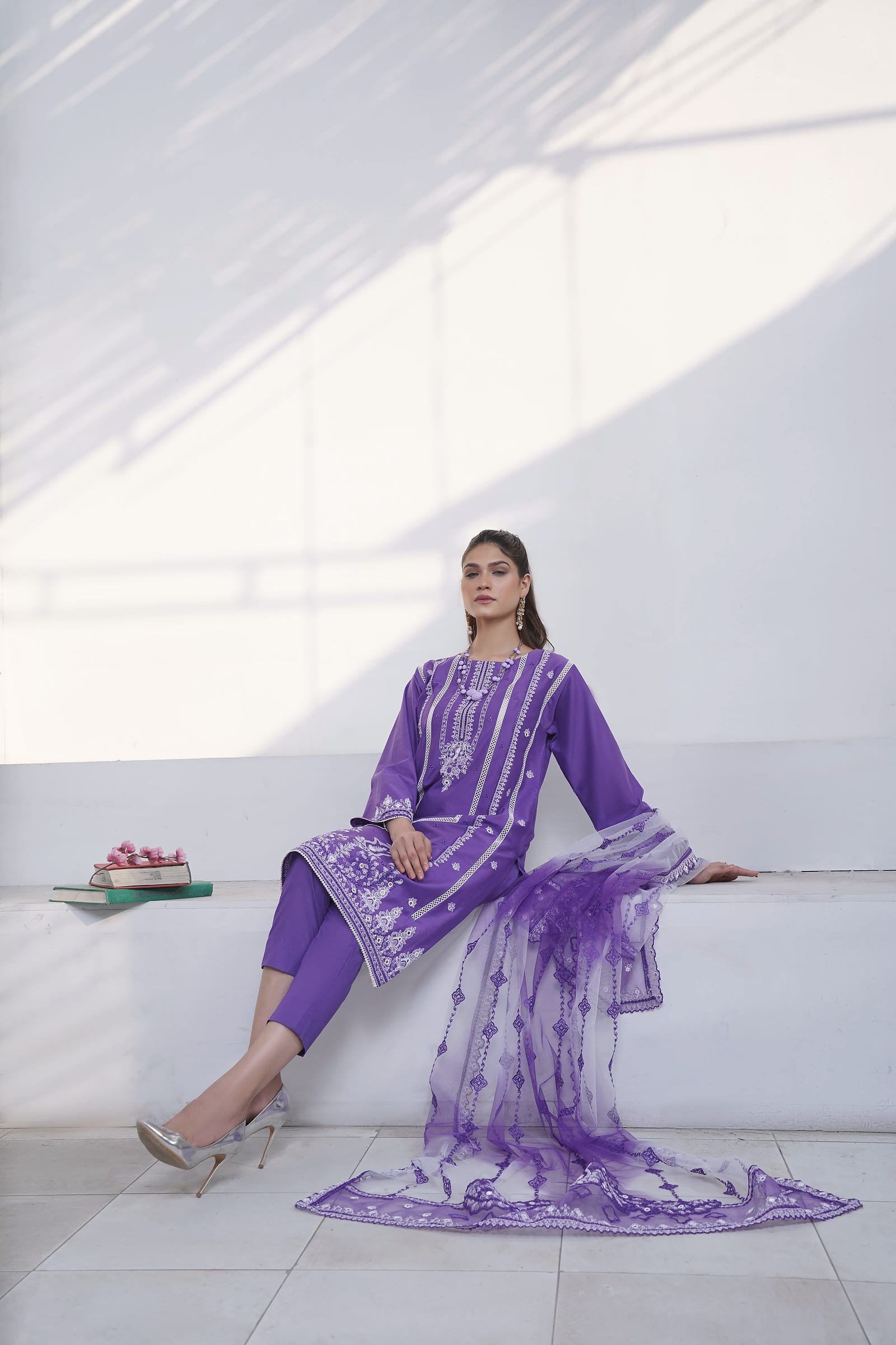 A woman stands full length in a white room, wearing a violet kurta set with white lace and cuff embroidery and a net dupatta.