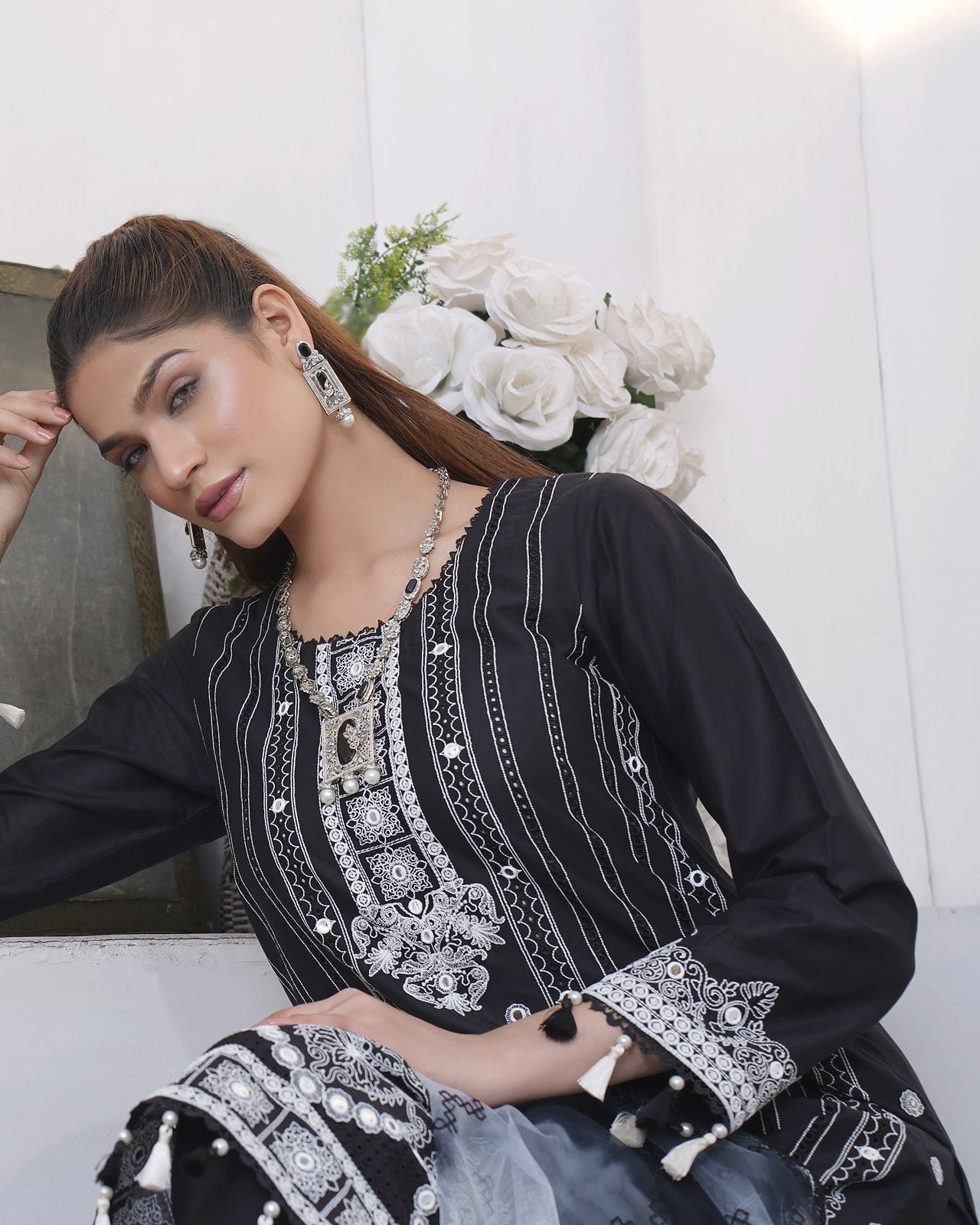A woman rests her head on a hand, with white roses behind, wearing a black kurta set with white stripe and mirror embroidery and a grey tassel dupatta.