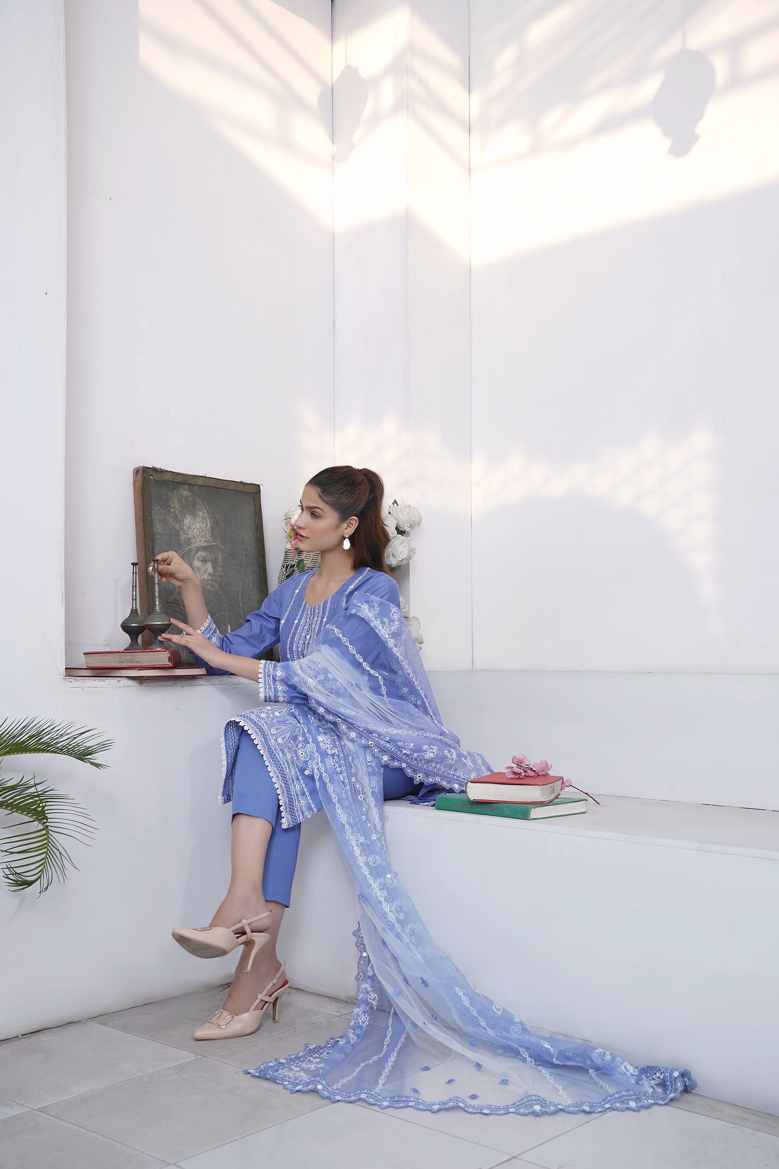 A woman sits in a white room, wearing a periwinkle kurta set with white embroidery and a white net dupatta.