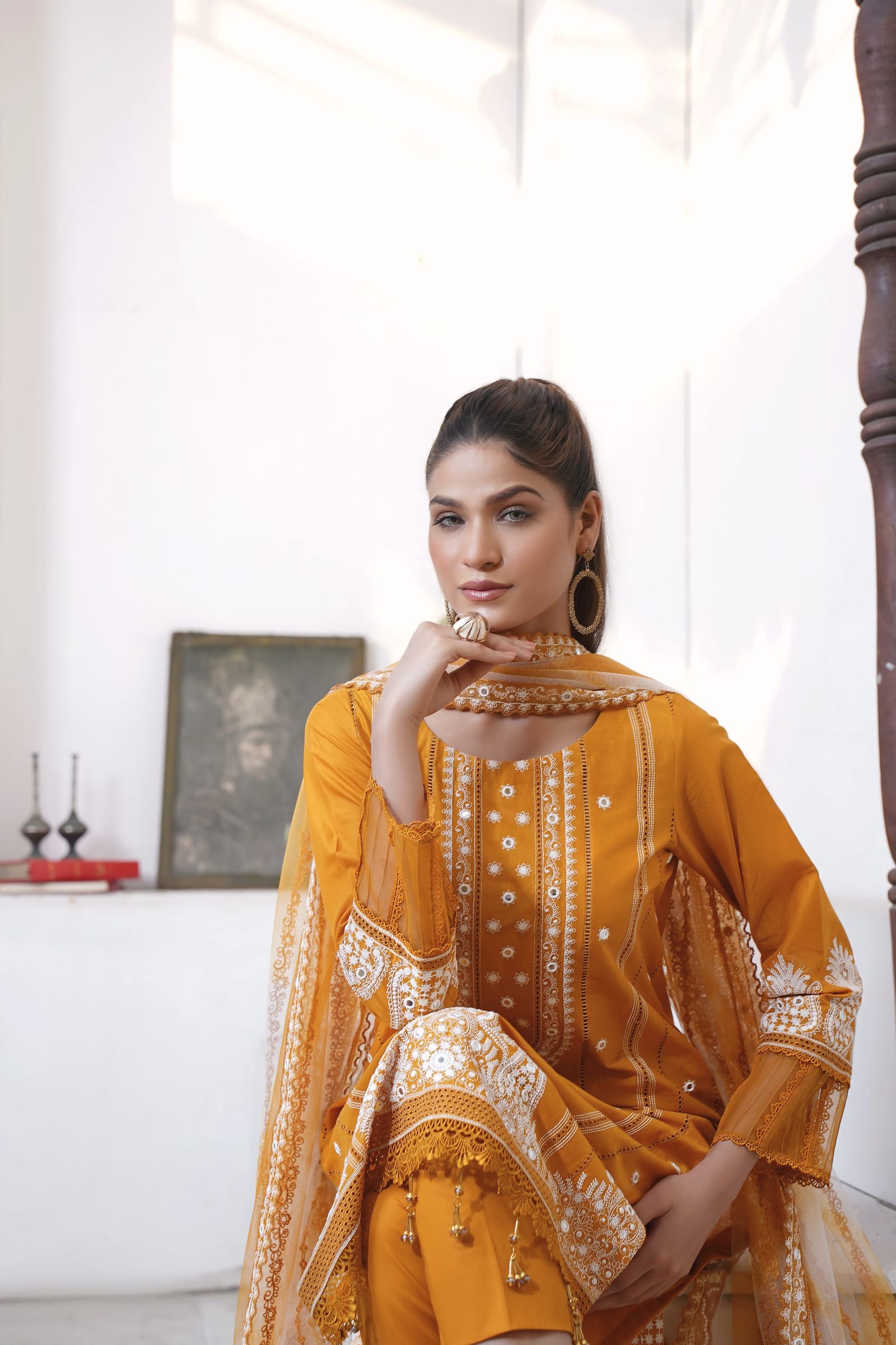 A woman is shown three-quarter length in a white room, wearing a mustard kurta set with white thread embroidery and a sheer net dupatta.