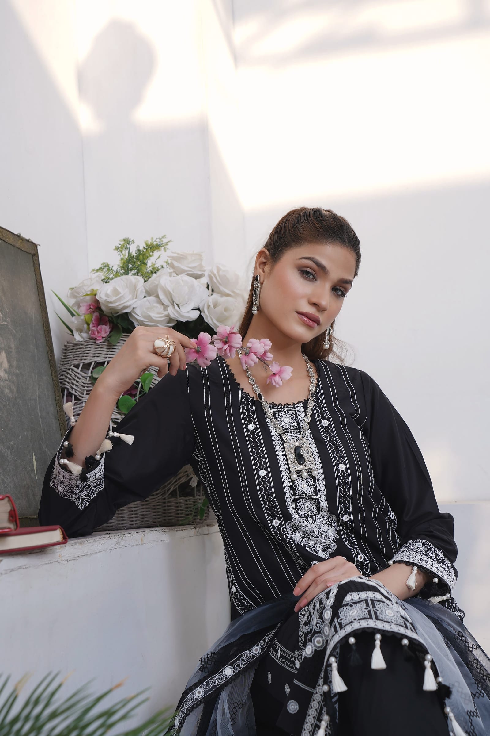 A woman is close in front of white roses, wearing a black kurta set with white stripe and mirror embroidery and a grey tassel dupatta.