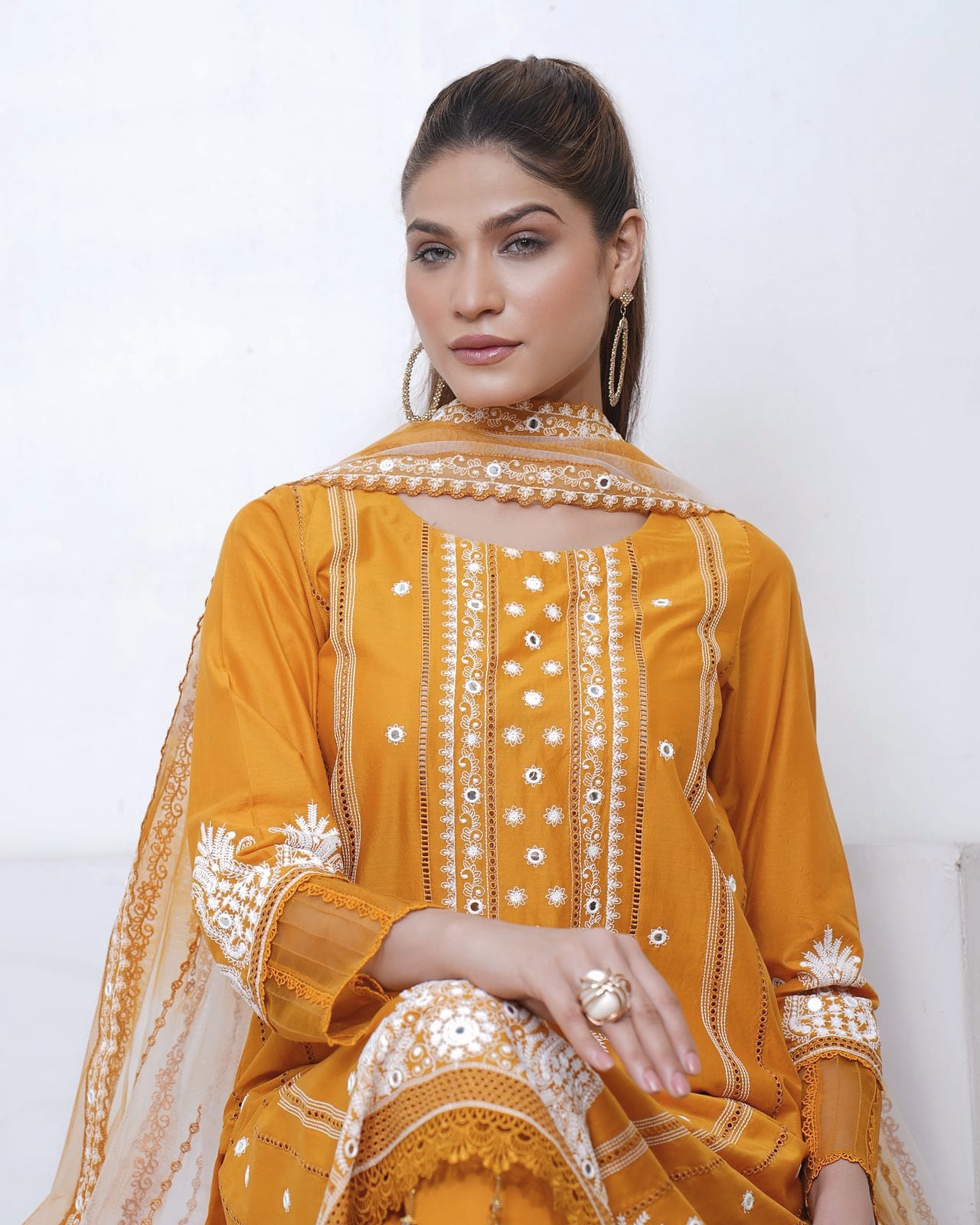 A woman sits in a white room, wearing a mustard kurta set with white thread embroidery and a sheer net dupatta.